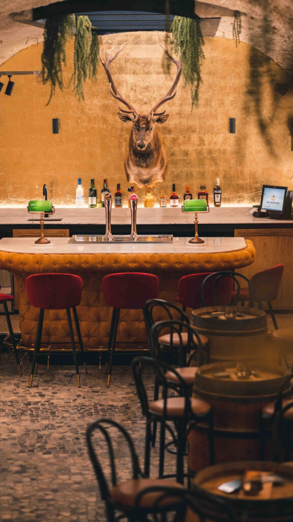 Gezellige barhoek in ons restaurant met een opgezette hertenkop als centraal kunststuk, omgeven door rode barkrukken en sfeervolle verlichting.