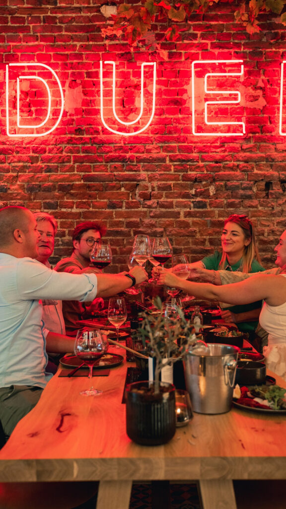 Vrienden proosten met wijn rond een tafel vol fonduepotten in ons sfeervol restaurant met opvallende 'FONDUE' neonlamp op een bakstenen muur.